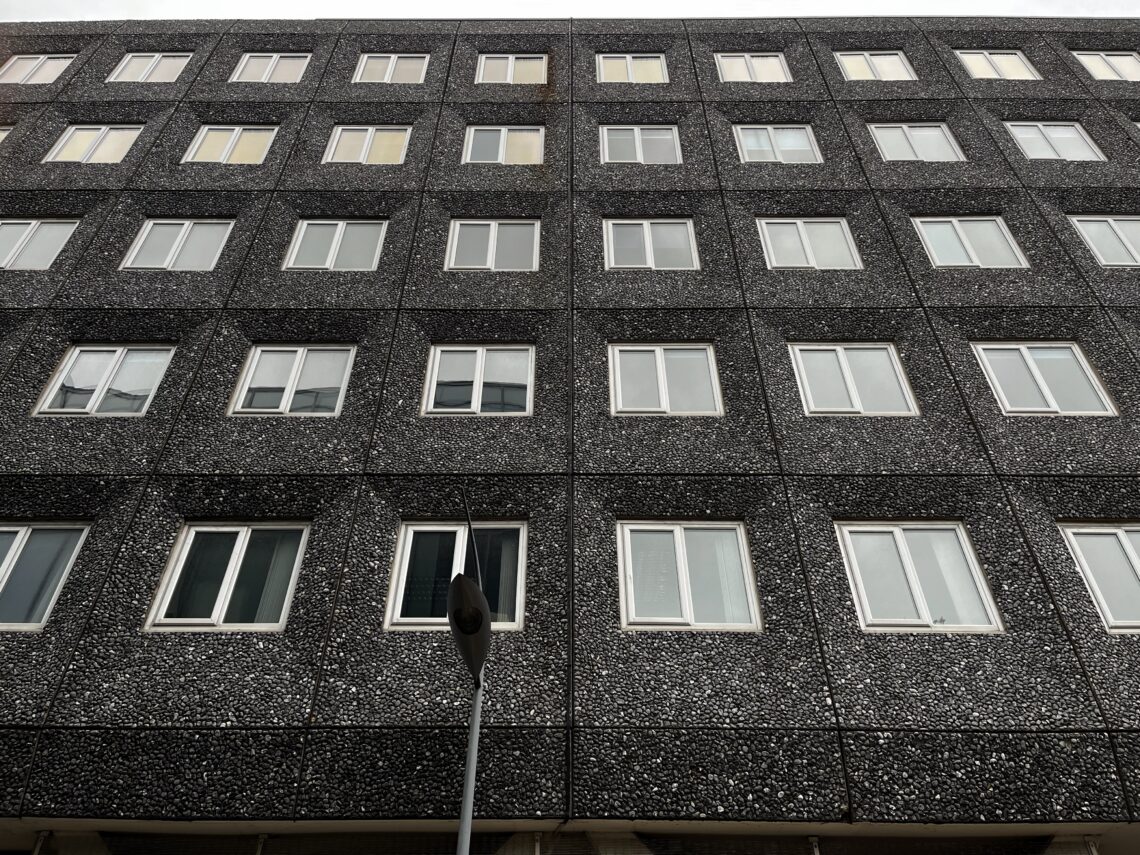 A modern building facade with a grid of square windows set in dark, speckled panels. There are six rows of windows, and a streetlamp is visible in the foreground centred near the lower part of the image.