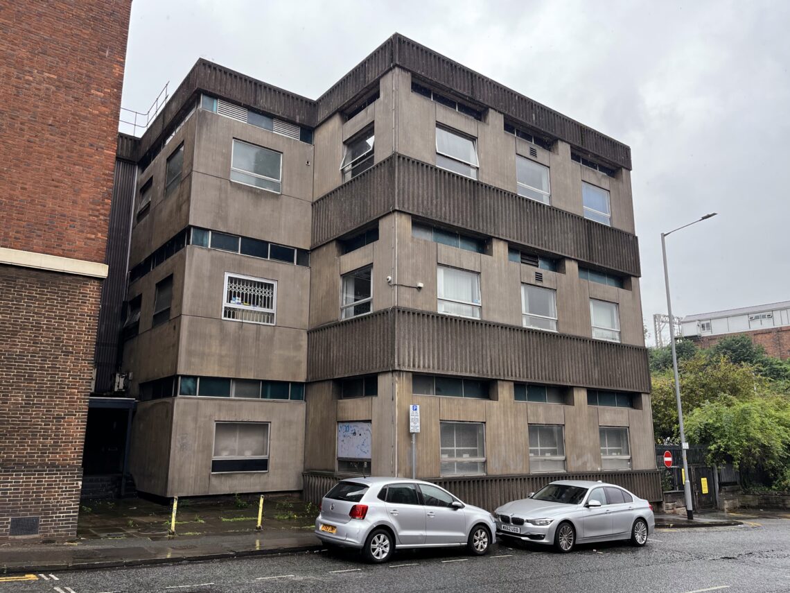 A grey, three-storey, concrete building with small, rectangular windows arranged in rows. Two cars, a silver hatchback and a silver saloon, are parked in front. The scene is overcast and wet, with patches of greenery to the right.