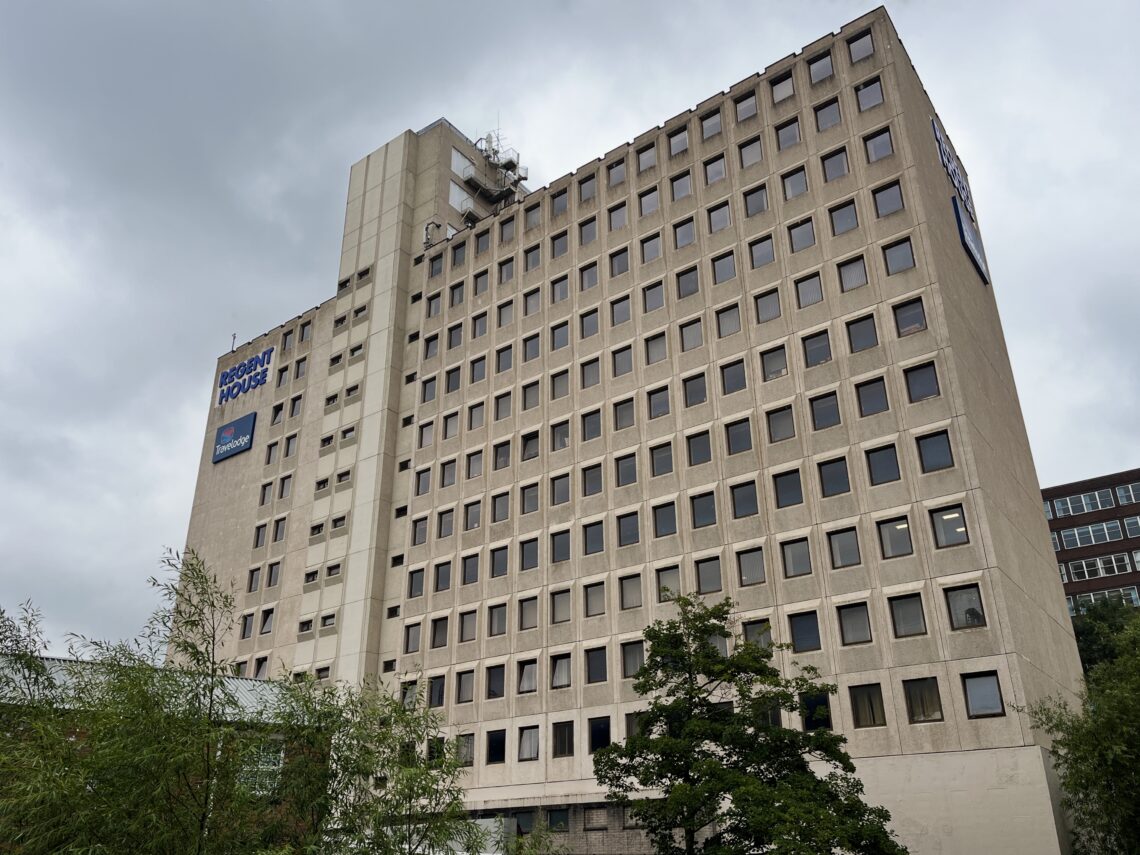 A tall beige building with many square windows, labelled “Regent House” with a blue sign near the top, stands under a cloudy sky. There are trees in the foreground and part of another building visible to the right.