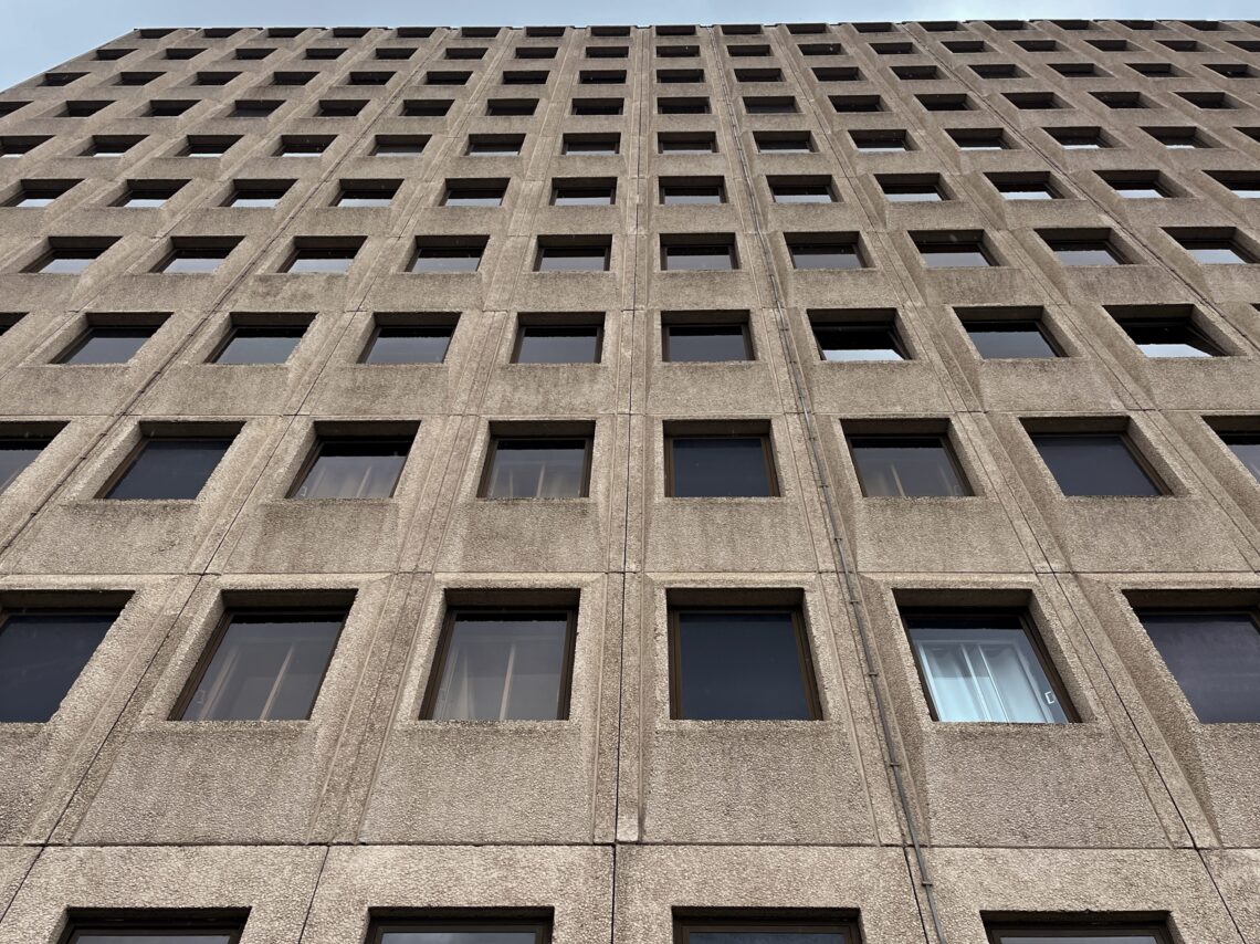 A close-up, upward-facing view of a tall, grey concrete building with many evenly spaced square windows arranged in a grid pattern. Most windows are dark, but one near the lower right corner has sheer white curtains. The sky is faintly visible at the top.