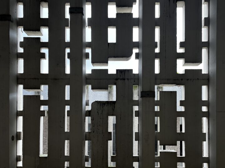 A concrete wall with a grid of geometric cut-outs in various shapes lets light and glimpses of sky through, casting shadows and creating a pattern of rectangles and squares across the surface.