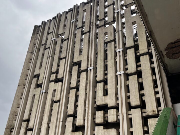 A close-up view of a tall, brutalist concrete building with vertical columns and geometric patterns. The weathered façade features deep grooves, angular cut-outs, and repeating rectangular forms. The sky above is overcast, adding a sombre tone.