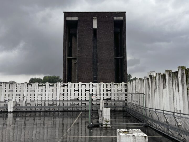 A wet, empty rooftop car park under a cloudy sky features a tall, dark brick tower in the centre, surrounded by white, weathered concrete barriers and posts. The scene looks desolate and industrial, with reflections visible on the rain-soaked ground.