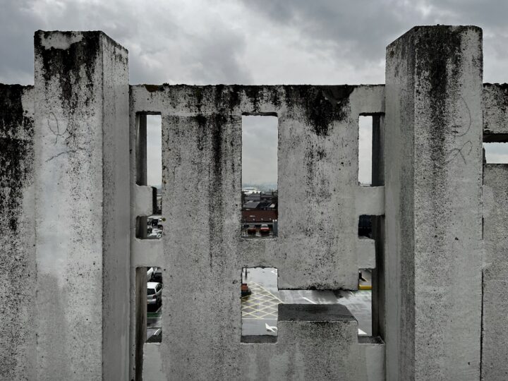 A heavily weathered concrete barrier with rectangular cut-outs stands on a rooftop. Through the central cut-out, a car park and distant buildings are visible under a cloudy grey sky, creating a sense of urban decay and overcast atmosphere.