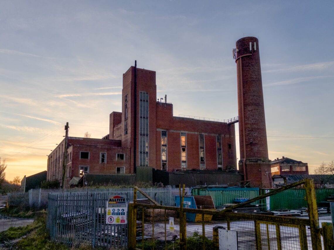 A large, abandoned red-brick industrial building with a tall cylindrical chimney stands behind a metal fence. The structure shows signs of decay. The sky is clear with a faint sunset. Warning signs are posted on the fence in the foreground.