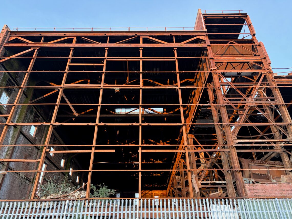 An old, rusted industrial steel structure with exposed beams and partially collapsed sections stands against a blue sky. A metal fence is in the foreground, and overgrown plants are visible inside the building’s lower level.
