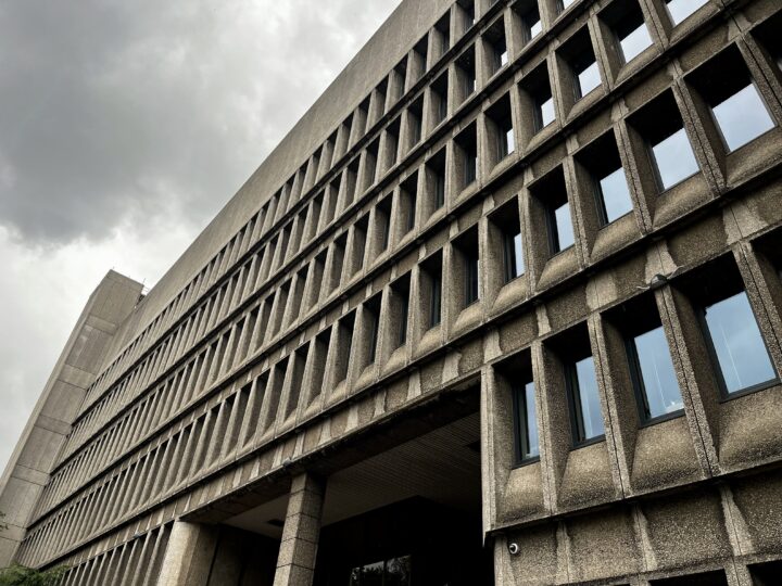 A large, concrete brutalist building with many rectangular windows is shown on a cloudy day. The photo is taken from a low angle, highlighting the repetitive geometric design and textured façade of the structure.