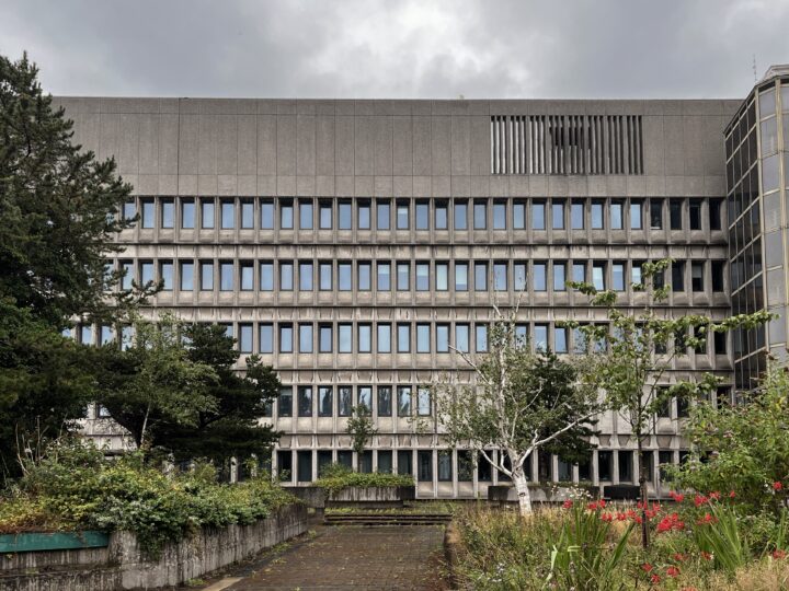 A large, grey, concrete office building with many rows of small rectangular windows, surrounded by overgrown plants, bushes, and trees under a cloudy, grey sky. The foreground includes a paved walkway and neglected landscaping.