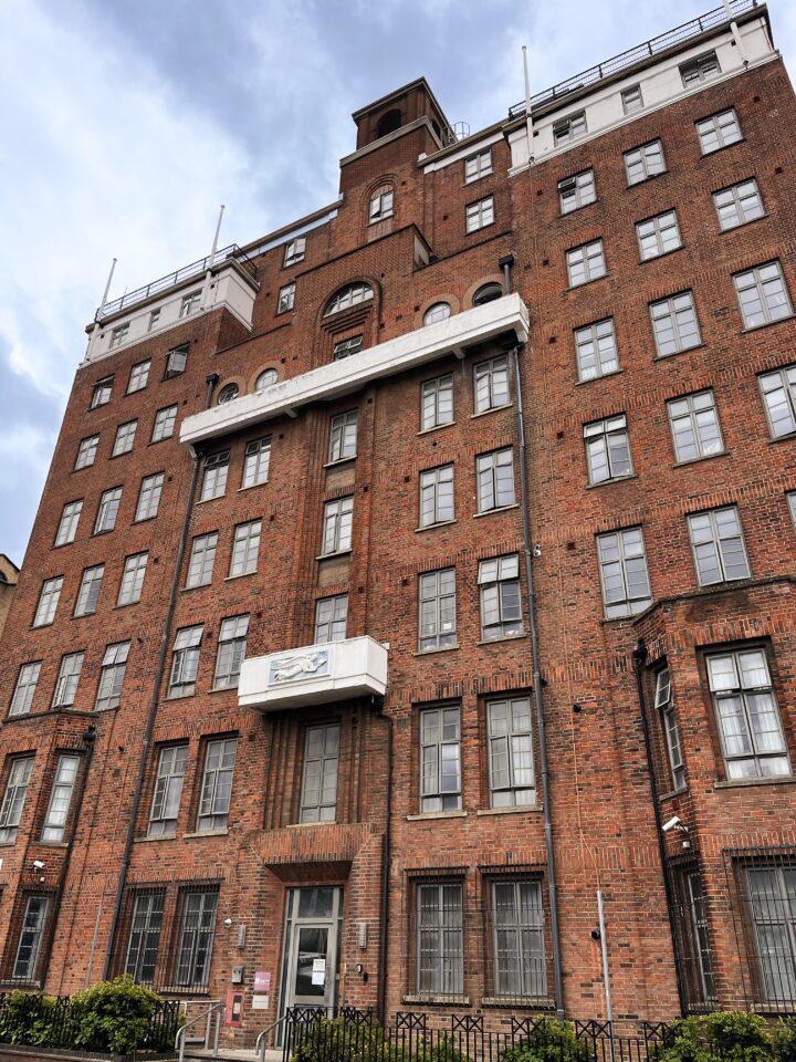 A tall red brick building with numerous grid-like windows, white trim, and two projecting white balconies. The sky above is cloudy, and a black railing and small green bushes are visible at ground level in front.