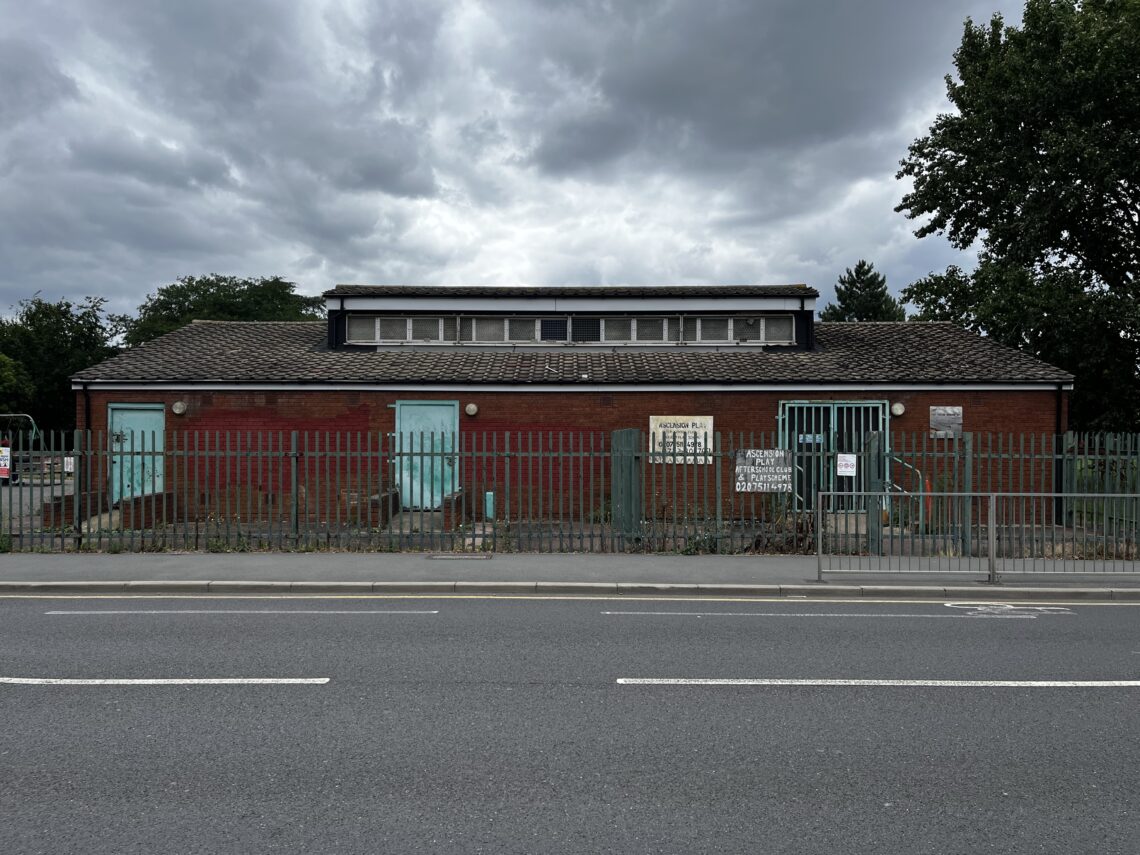 A small, single-storey red brick building with a sloping roof sits behind a tall metal fence. There are two turquoise doors and signs on the front, with overcast skies and trees in the background. The building faces a quiet, empty road.