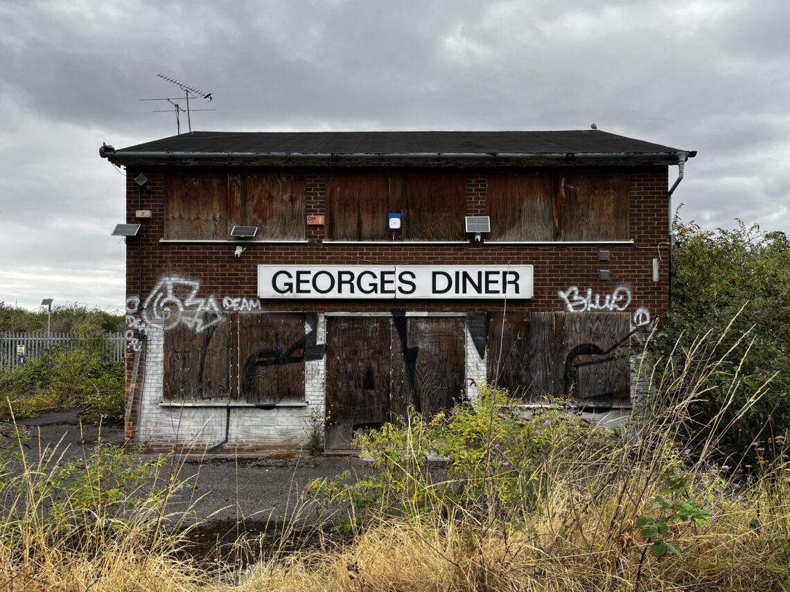 A boarded-up, abandoned brick building with a large sign reading GEORGE'S DINER. The windows and doors are covered with timber, graffiti is visible on the walls, and overgrown weeds dominate the foreground. The sky is overcast.