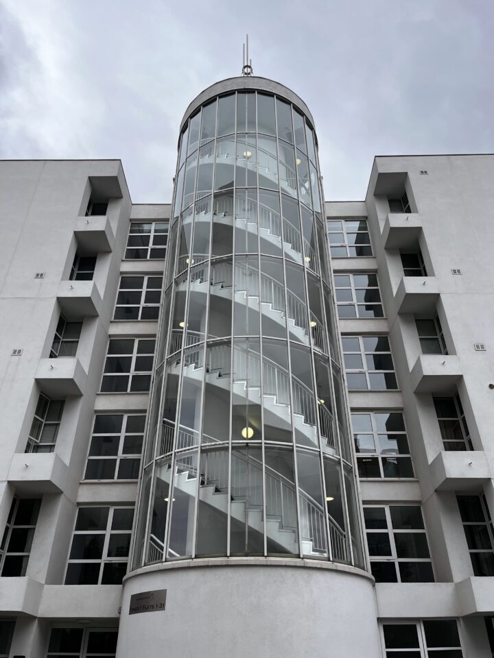 A modern white building with a central cylindrical glass tower enclosing a spiral staircase. The building has symmetrical rectangular windows and balconies on both sides. The sky above is overcast with grey clouds.