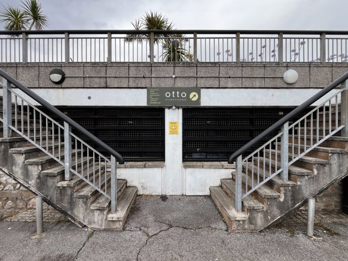 A closed shopfront with a sign reading otto. Two symmetrical staircases lead up on either side, separated by a central railing and a grated window. Overcast sky and some palm trees are visible above the structure. The area appears empty.