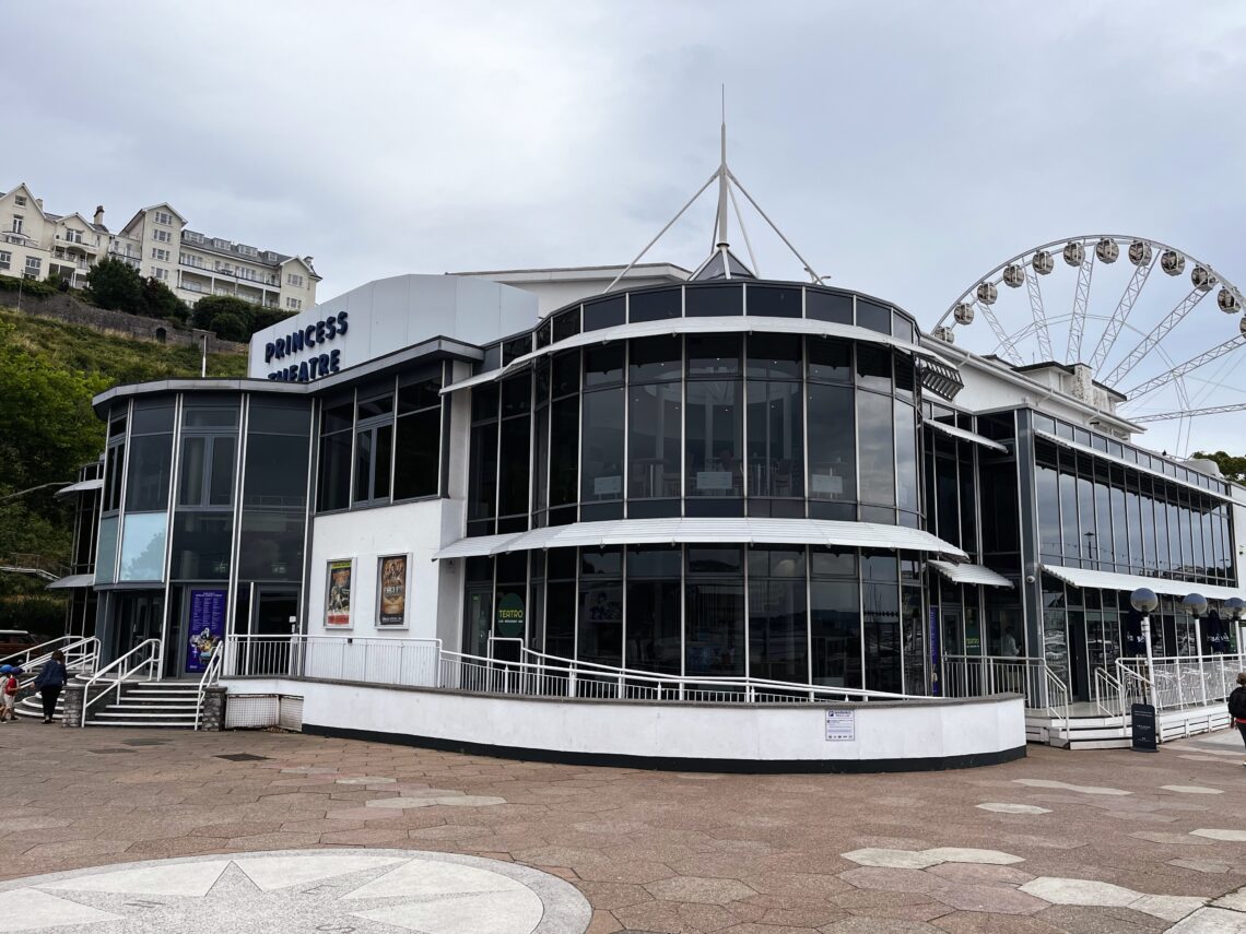 A modern glass-fronted building labelled Princess Theatre with large rounded windows, white trim, and ramp access. Posters are visible near the entrance. In the background, a Ferris wheel and hillside buildings can be seen under a cloudy sky.