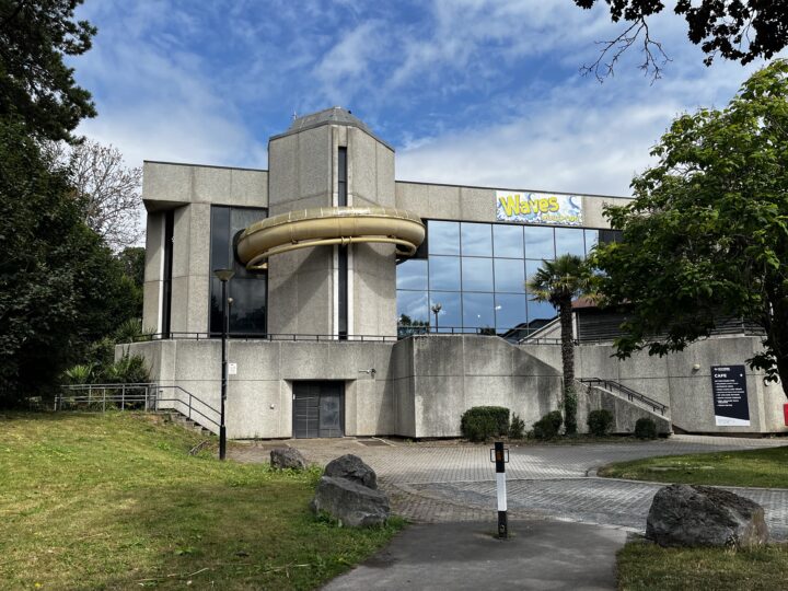 A modern, grey concrete leisure centre with large mirrored windows reflects the sky. A yellow Waves sign is on the upper wall. Bushes and palm trees surround the entrance. A paved path and grassy area with rocks are in front, under a partly cloudy sky.