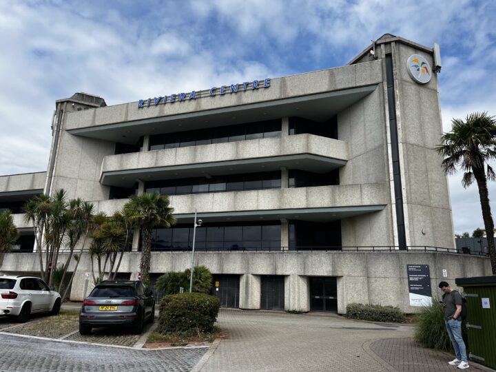 A large, modern concrete building labelled “RIVIERA CENTRE” has balconies and palm trees in front. Two parked cars and two people standing near a sign are visible. The sky is partly cloudy, and the area looks clean and well-maintained.
