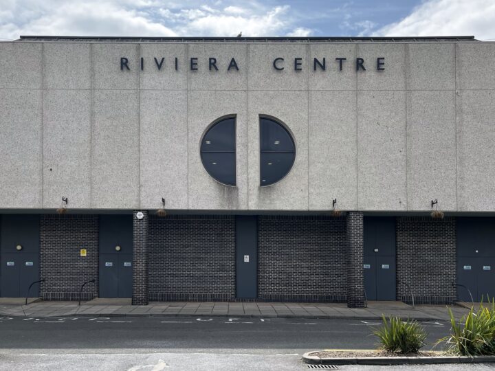 A grey, concrete building with RIVIERA CENTRE in bold letters at the top. A large circular window is above three blue doors. The lower facade features dark brickwork. Some plants and bicycle racks are visible near the pavement in the foreground.