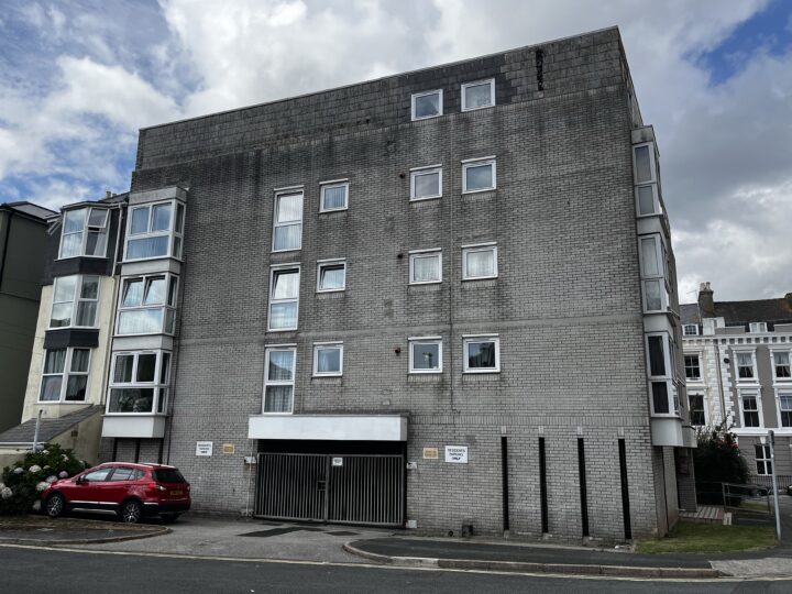 A large, grey, four-storey block of flats with many rectangular windows. The façade is plain and made of concrete bricks. A car park is visible below, with a red car parked on the left. The sky is cloudy and other buildings are partly visible nearby.