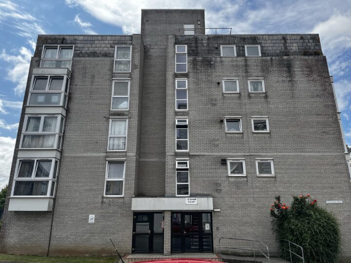 A five-storey, grey brick apartment building with large rectangular windows, some protruding in box-like frames. Double glass doors mark the entrance, with a sign reading “Osney Court” above. A bush with orange flowers grows on the right, under a partly cloudy sky.