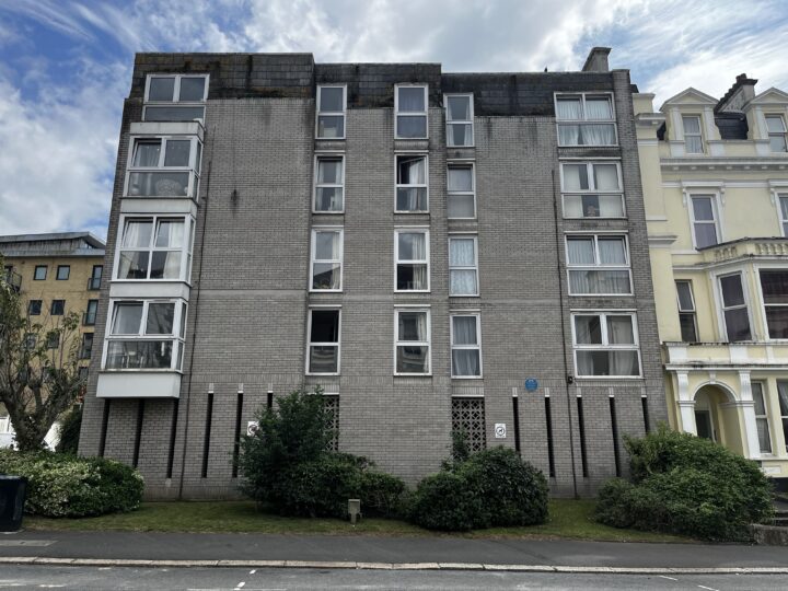 A five-storey grey brick block of flats with large pane windows, some open. Bushes and small shrubs line the base. The building sits between a pale yellow house and another structure. A blue plaque is visible on the wall, and the sky is partly cloudy.