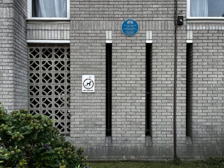 A grey brick building with vertical slits, a blue English Heritage plaque above, and a No Parking sign on the wall. Decorative concrete blocks are to the left, a bush with green leaves in the foreground, and windows near the top.
