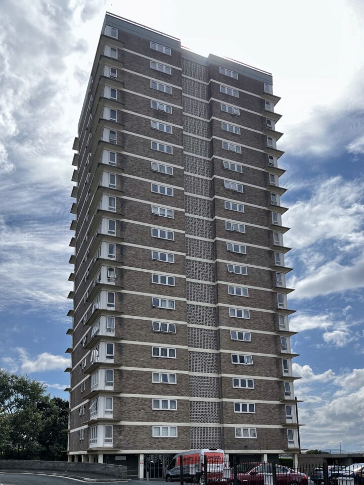 Tall, modern block of flats with brown brick exterior and white window frames. The block has around 15 storeys and is set against a partly cloudy, sunny sky. Cars, including a red van, are parked at the base near a black metal fence.