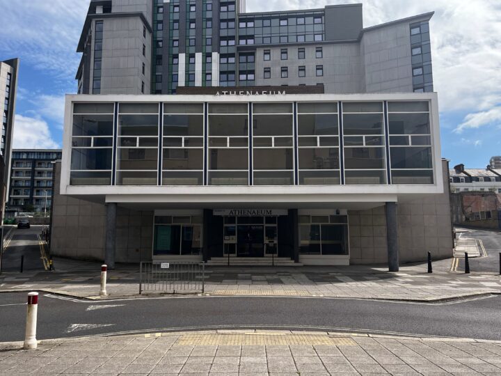 A modern, rectangular building labelled “ATHENAEUM” features large glass windows on the upper floor and a sheltered entrance below. The facade is grey, with bollards, a ramp, and a pedestrian crossing visible in front on a sunny day.