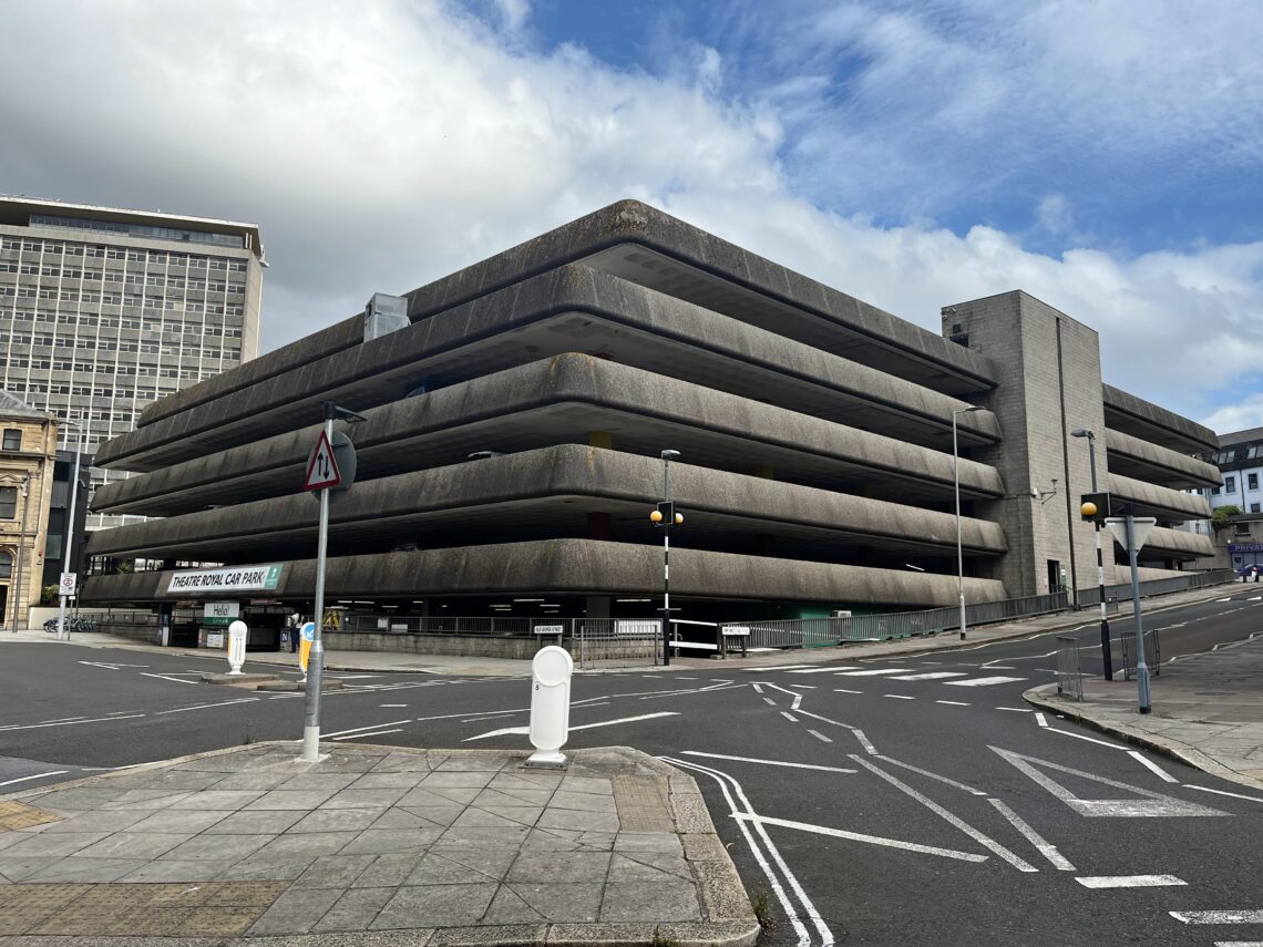 A large, multi-storey concrete car park with rounded edges and horizontal lines dominates a mostly empty urban street. The sky is partly cloudy, and a tall office building is visible in the background. Road signs and pedestrian crossings surround the car park.