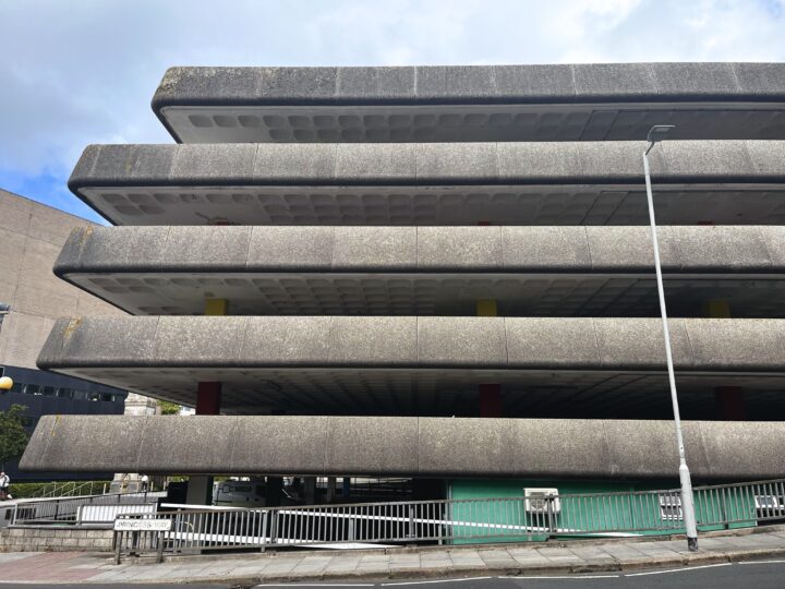 A multi-storey concrete car park with wide, flat, textured floors and gaps between levels. A white streetlamp stands in front, and a metal fence borders the pavement. Yellow and red pillars are visible inside, with a partly cloudy sky above.