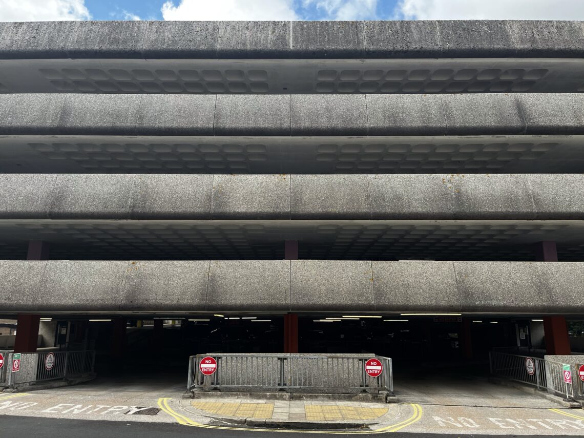 A multi-storey concrete car park with three visible levels. No Entry signs and barriers block the entrance. Faded NO ENTRY is painted on the ground. The sky is partly cloudy above the building. The scene appears quiet and empty.