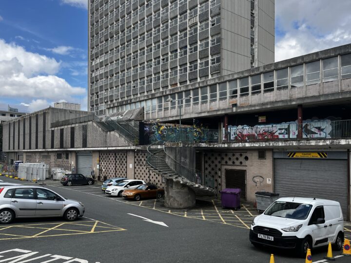 A grey, concrete car park by a brutalist tower block. Several cars are parked below. A spiral staircase leads to a walkway covered in colourful graffiti. The scene is urban, with cloudy blue sky above and yellow road markings on the ground.
