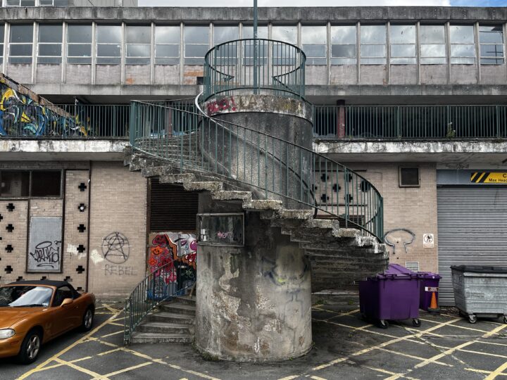 A concrete spiral staircase with a green metal railing wraps around a cylindrical pillar in front of a graffiti-covered, urban building. Yellow parking lines, a parked orange car, purple bins, and industrial details are visible in the scene.
