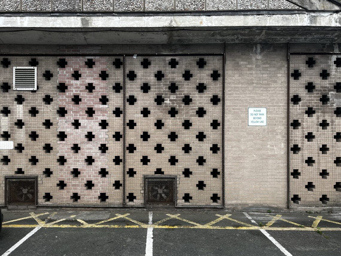 A brick wall with a grid of cross-shaped cut-outs, two vents near the bottom, and a white sign reading “Please do not park in front of roller bar.” Yellow diagonal parking lines and white lines mark the ground in front.