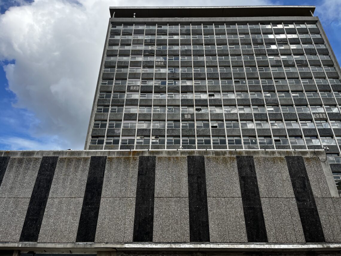 A tall, rectangular, concrete building with many small, dirty windows stands behind a shorter structure with alternating vertical dark and light stripes. The sky is partly cloudy, with blue sky visible on the left and a large white cloud on the right.