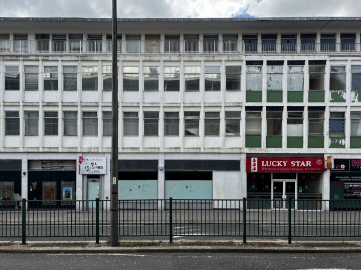 A rundown, multi-storey white building with many boarded-up windows. On the ground floor are the closed “LUCKY STAR” Chinese restaurant, a sign for “EXPRESS” convenience shop, and more vacant shopfronts. A metal fence runs along the road in front.