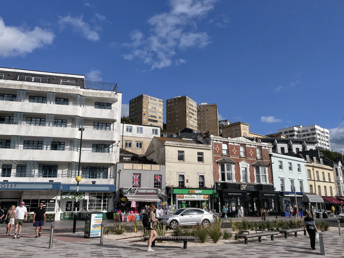 A sunny street scene features people walking past colourful shops and cafés on the ground floor of older buildings. Modern blocks of flats rise in the background under a bright blue sky with scattered clouds. A silver car is parked near the pavement.