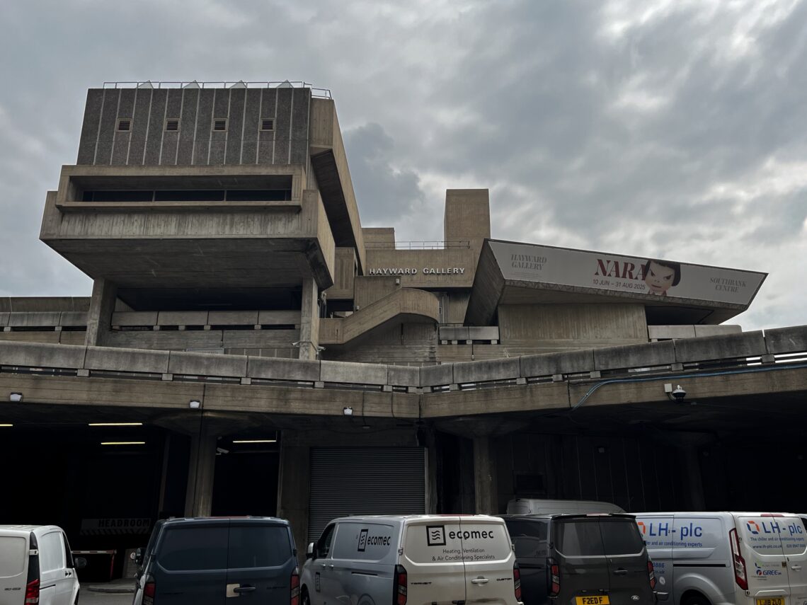 A brutalist concrete building with angular shapes houses the Hayward Gallery. A sign above a ramp reads Hayward Gallery. A large billboard advertises a NARA exhibition. Several white vans are parked below; the sky is overcast.