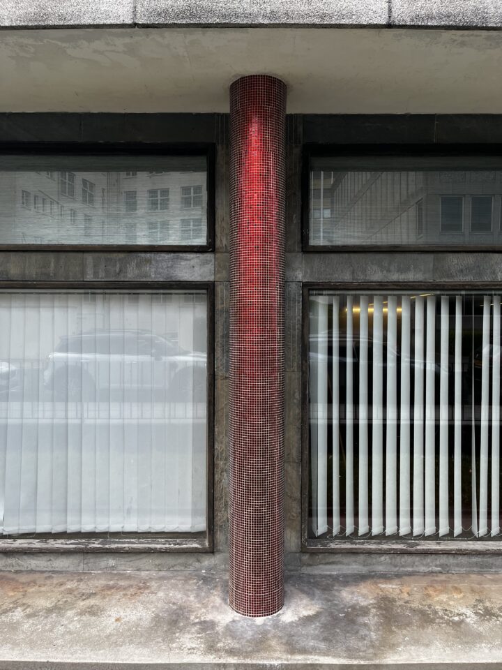 A cylindrical column covered in small red tiles stands between two large windows with vertical white blinds. The column casts a slight shadow, and reflections of buildings and cars are visible in the glass behind it. The scene appears urban and modern.