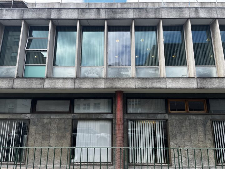 A concrete building facade with tall vertical windows, some with closed blinds and one open window. The lower level has grey stone and vertical bars. Reflections of clouds and city buildings appear in the glass. A single red pillar stands in the centre.
