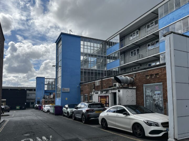 A narrow street with parked cars runs beside a blue and white multi-storey building with glass stairwells. The brick ground floor has graffiti and industrial vents. Cloudy sky and scattered rubbish bins are visible in the background.