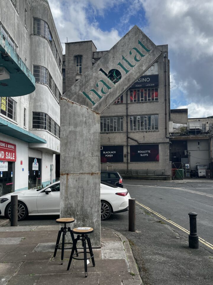 A concrete sign reading “habitat” is bent at a sharp angle on a city street. Two wooden bar stools sit in front of it. Behind, there are old grey buildings, a parked white car, and signs for a casino, all under a partly cloudy sky.