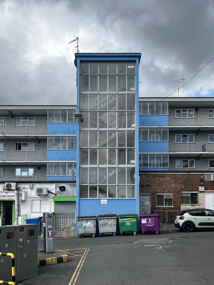 A blue block of flats with a glass-enclosed stairwell in the centre. Three storeys are visible. In front are several skips and bins. To the right is a white car, and the sky above is mostly cloudy with patches of sunlight.