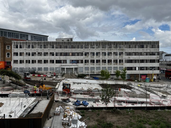 A large, weathered, grey building with many windows stands behind a construction site. Workers, machinery, and building materials are scattered around the site. Some trees and patches of earth are visible. The sky above is cloudy with patches of blue.