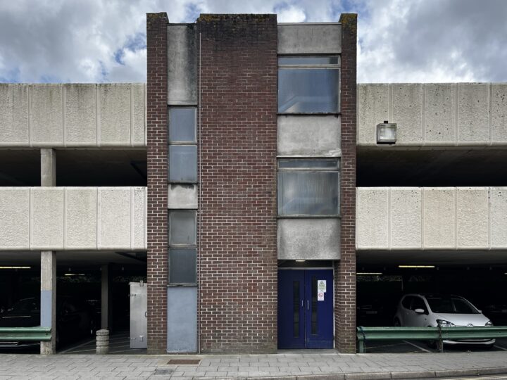 A symmetrical section of a multi-storey car park features a central brick stairwell with windows and a blue door, flanked by open concrete parking bays. Several parked cars and a cloudy sky are visible. The setting appears urban and utilitarian.