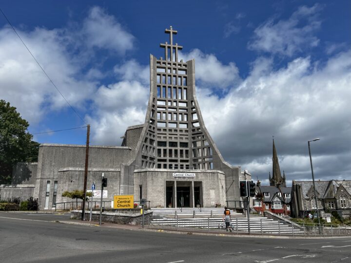 A modern concrete church with a large grid-like tower and central cross stands at a street corner. Steps lead to the entrance labelled Central Church. A person in an orange top walks nearby. Older stone buildings and a church spire are visible in the background.