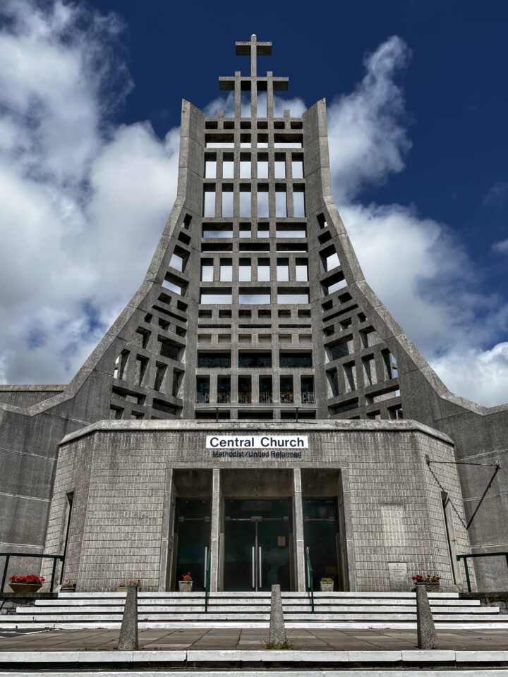 A modern concrete church with a tall, geometric façade featuring grids and a large cross at the top. The entrance has glass doors and a sign reading “Central Church.” The sky is blue with scattered clouds. Potted flowers decorate the entrance area.