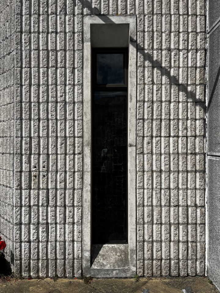 A tall, narrow window set in a textured, grey concrete block wall. The window is framed with a simple concrete border and casts a shadow. Light and shadow highlight the rough, grid-like pattern of the blocks. The ground below is worn and partially shaded.