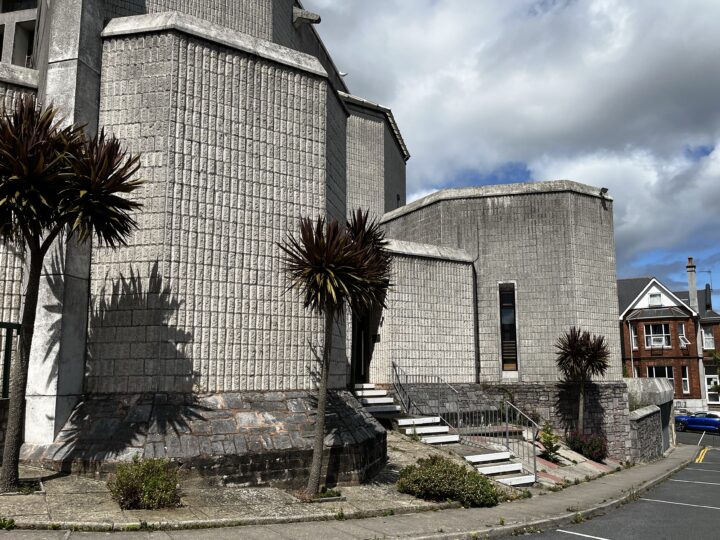 A brutalist building with textured concrete walls, narrow vertical windows, and angular forms stands under a partly cloudy sky. Two spiky-leaved palm trees cast shadows on the facade, and steps lead up to the main entrance from a sloped pavement.