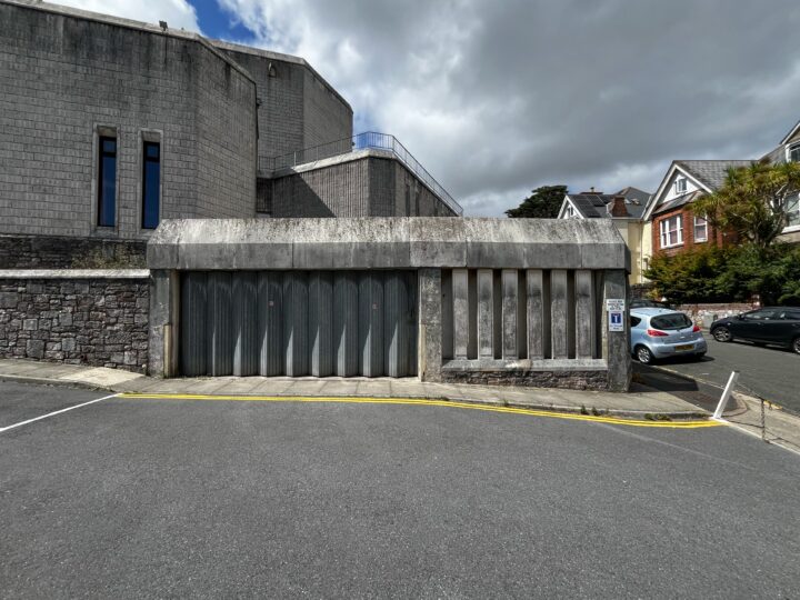 A concrete garage with vertical metal doors is set into a stone building. Yellow hazard tape blocks the entrance, and there are parked cars and residential houses to the right. The sky is partly cloudy.