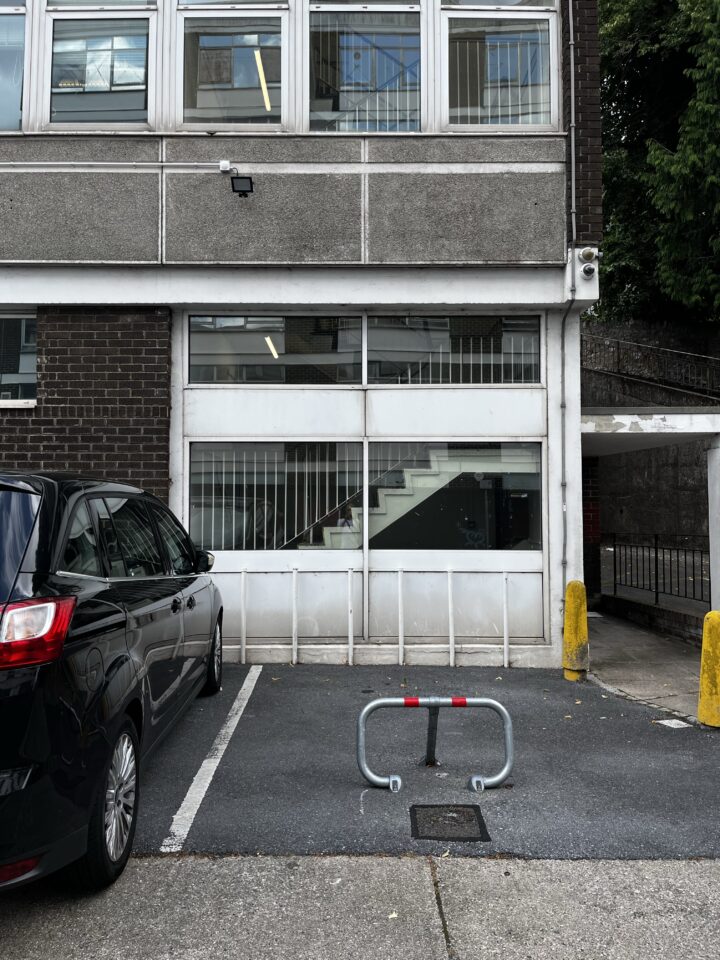 A black car is parked beside an empty parking space with a metal barrier. Behind them is a building with large windows, visible stairs inside, grey brick and concrete walls, and a CCTV camera above the middle window. Trees are on the right edge.